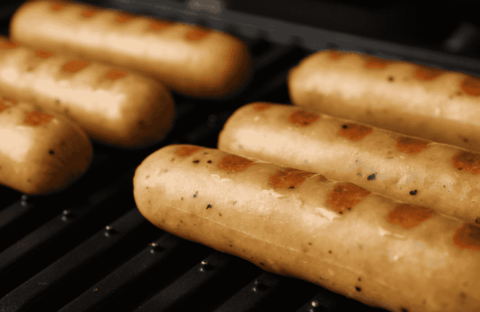 Six sausages with visible grill marks are cooking on a black grill. The sausages are light brown and evenly spaced, with a close-up focus highlighting their texture and the heat of the grill.