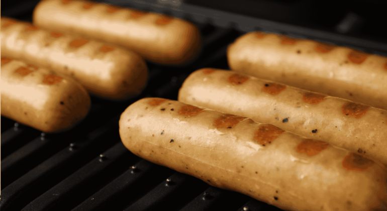 Six sausages with visible grill marks are cooking on a black grill. The sausages are light brown and evenly spaced, with a close-up focus highlighting their texture and the heat of the grill.