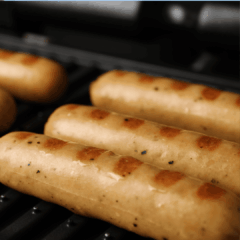 Four sausages grilling on a barbecue, with visible grill marks and slight browning on the surface. The background is slightly out of focus, emphasizing the sausages in the foreground.