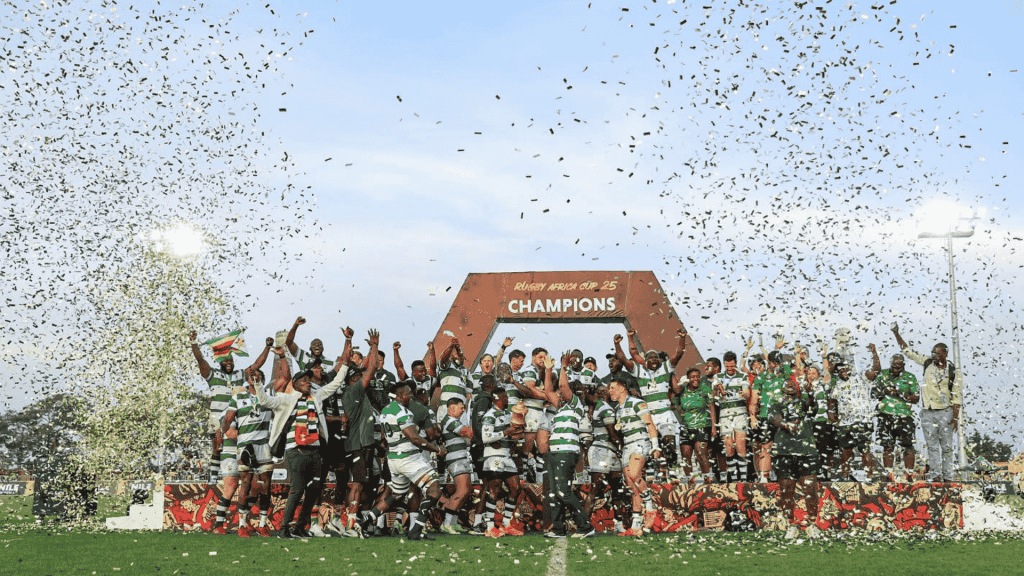 A rugby team celebrates winning a championship on a field, standing under a large CHAMPIONS sign while confetti falls around them. Players and coaches raise their hands in victory and joy.