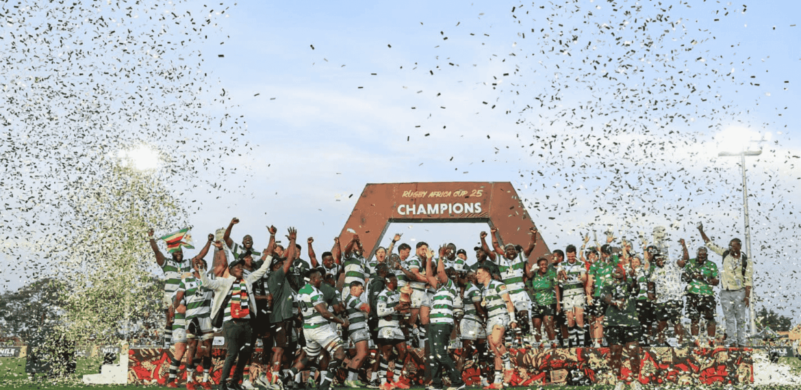 A rugby team celebrates winning a championship on a field, standing under a large CHAMPIONS sign while confetti falls around them. Players and coaches raise their hands in victory and joy.