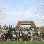 A rugby team celebrates winning a championship on a field, standing under a large CHAMPIONS sign while confetti falls around them. Players and coaches raise their hands in victory and joy.
