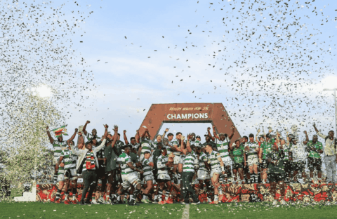 A rugby team celebrates winning a championship on a field, standing under a large CHAMPIONS sign while confetti falls around them. Players and coaches raise their hands in victory and joy.