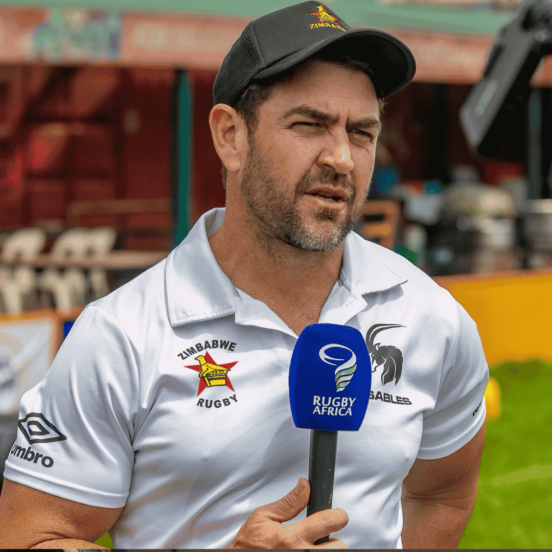 A man wearing a black cap and a white Zimbabwe Rugby polo shirt speaks into a blue Rugby Africa microphone during an outdoor interview.