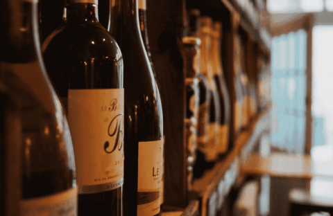 Close-up of wine bottles on a shelf in a dimly lit store or wine shop, with blurred shelves and a door visible in the background.