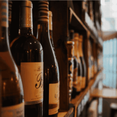 A close-up of wine bottles lined up on a wooden shelf in a dimly lit wine shop or cellar, with other bottles and shelves blurred in the background.
