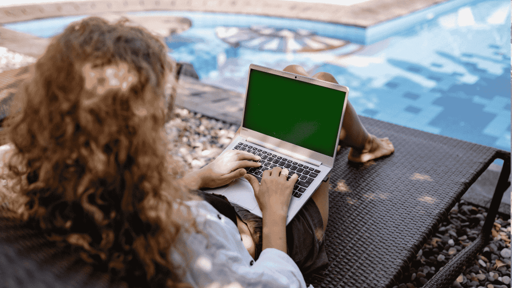 A person with curly hair sits on a lounge chair by a pool, using a laptop with a green screen. Sunlight casts shadows on the ground and water.