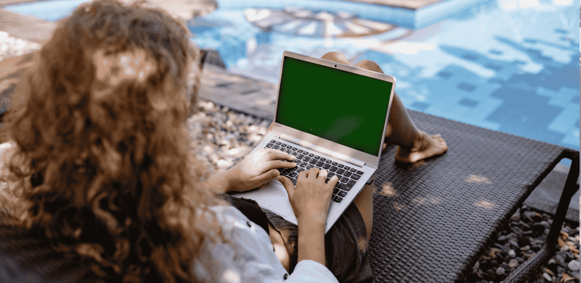 A person with curly hair sits on a lounge chair by a pool, using a laptop with a green screen. Sunlight casts shadows on the ground and water.