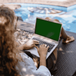 A person with curly hair sits on a lounge chair by a pool, using a laptop with a green screen. Sunlight casts shadows on the ground and water.