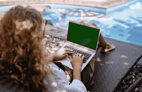 A person with curly hair sits on a lounge chair by a pool, using a laptop with a green screen. Sunlight casts shadows on the ground and water.
