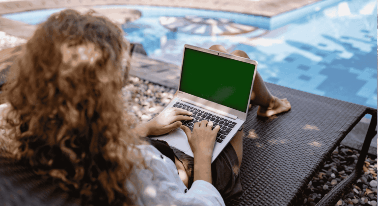 A person with curly hair sits on a lounge chair by a pool, using a laptop with a green screen. Sunlight casts shadows on the ground and water.