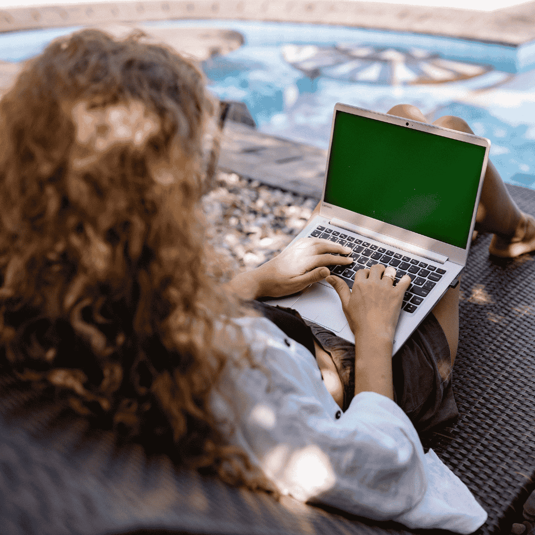 A person with curly hair sits on a lounge chair by a pool, working on a laptop with a green screen.
