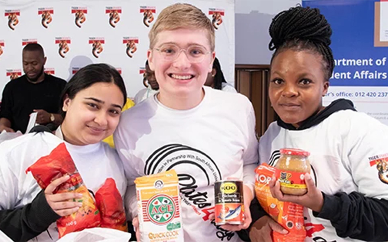 Three smiling people wearing white T-shirts hold various food items, including noodles, chips, and jars. A branded banner and other people are visible in the background.