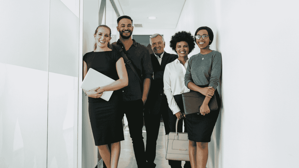 Five professionally dressed people stand smiling in a hallway, holding folders, bags, or laptops, suggesting a business or office environment.