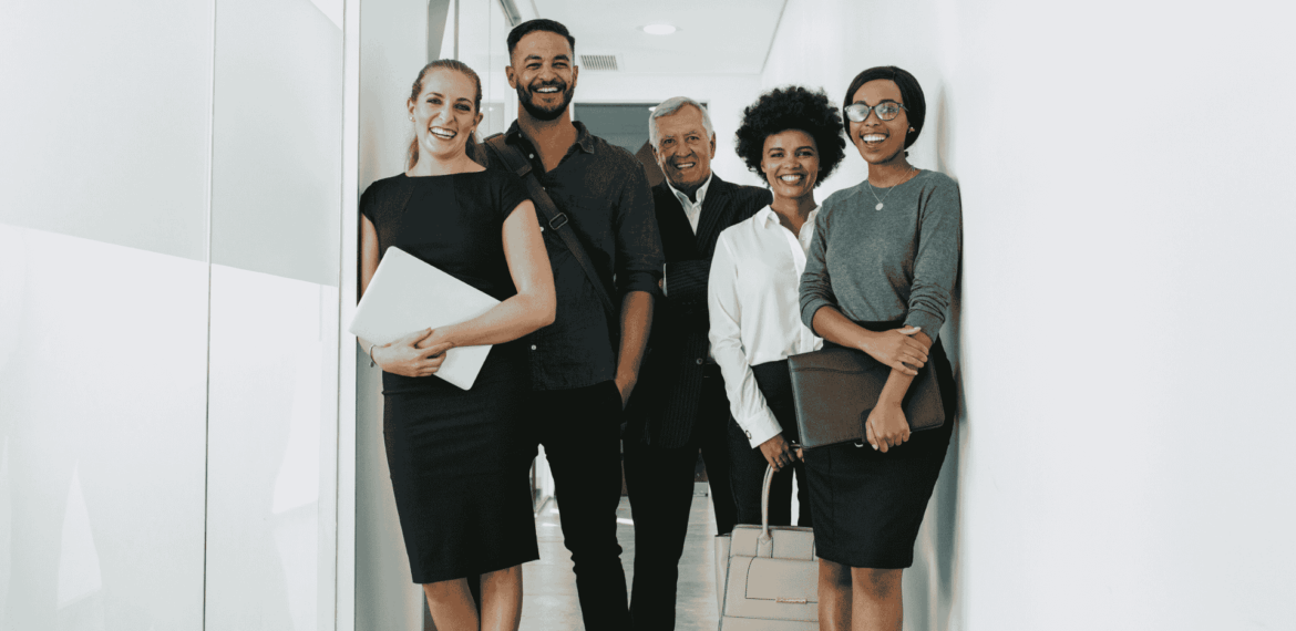 Five professionally dressed people stand smiling in a hallway, holding folders, bags, or laptops, suggesting a business or office environment.