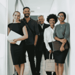 Five professionally dressed people stand smiling in a hallway, holding folders, bags, or laptops, suggesting a business or office environment.