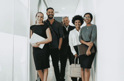 Five professionally dressed people stand smiling in a hallway, holding folders, bags, or laptops, suggesting a business or office environment.