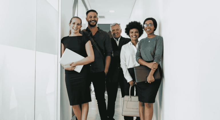 Five professionally dressed people stand smiling in a hallway, holding folders, bags, or laptops, suggesting a business or office environment.