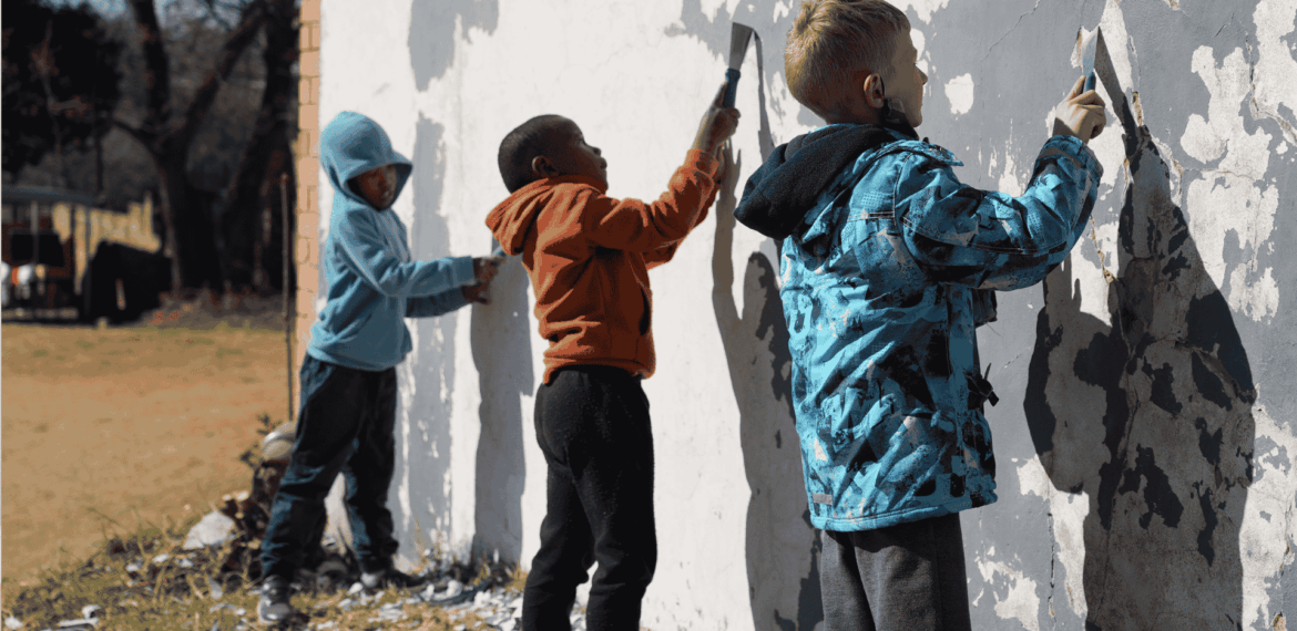 Three young children stand side by side, scraping old paint off a large outdoor wall with tools, focused on their work. They wear hooded jackets and casual pants, suggesting cool weather.