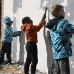 Three young children stand side by side, scraping old paint off a large outdoor wall with tools, focused on their work. They wear hooded jackets and casual pants, suggesting cool weather.