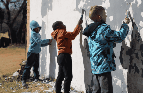 Three young children stand side by side, scraping old paint off a large outdoor wall with tools, focused on their work. They wear hooded jackets and casual pants, suggesting cool weather.