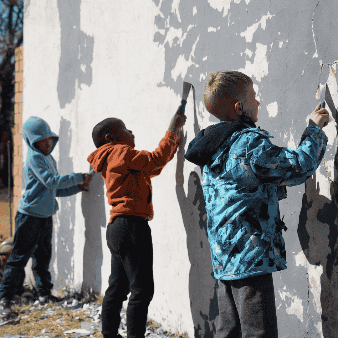 Three children peel old paint from an outdoor wall using scrapers on a sunny day. They wear jackets and hoodies, focused on removing the flaking paint together.