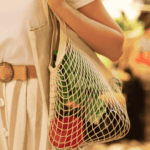 A person wearing a white shirt and striped pants carries a reusable net bag filled with produce while shopping in a grocery store.