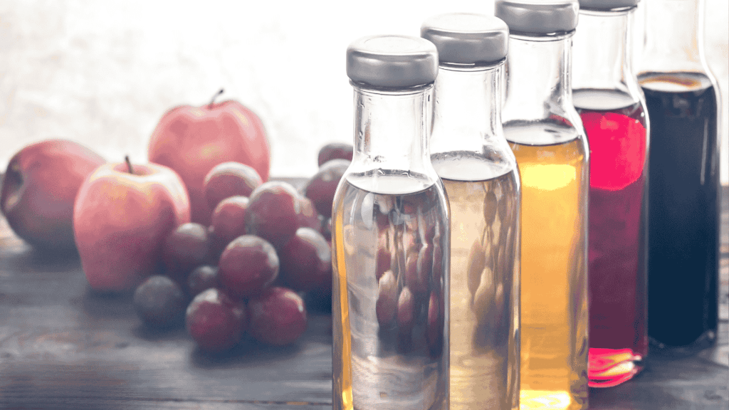 Four glass bottles filled with different colored liquids are lined up on a wooden table, with apples and grapes blurred in the background.