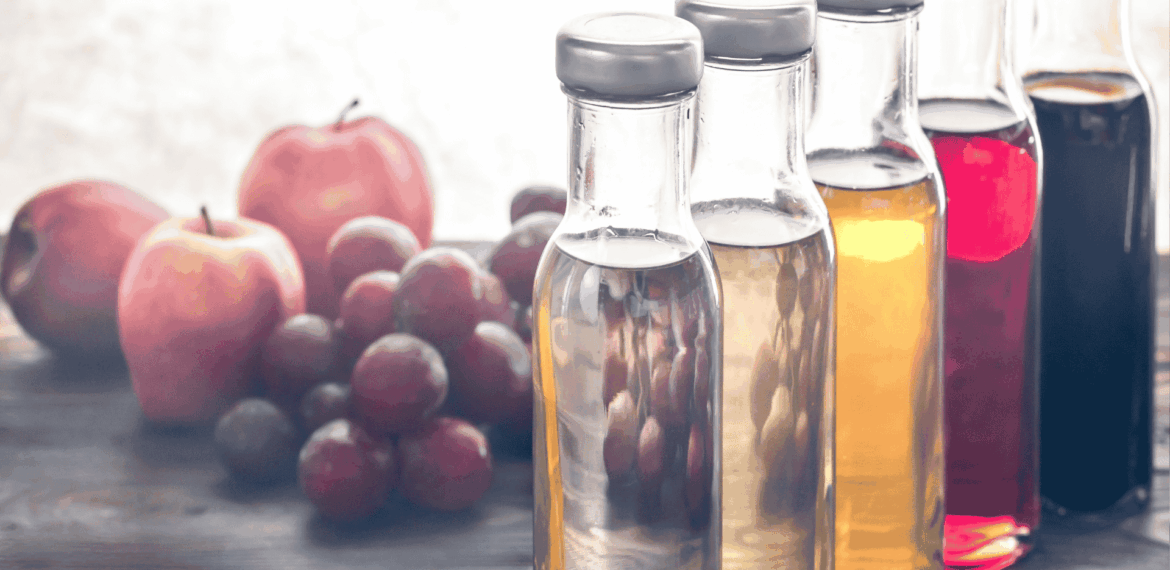 Four glass bottles filled with different colored liquids are lined up on a wooden table, with apples and grapes blurred in the background.
