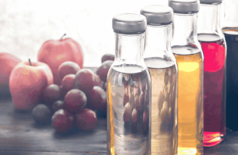 Four glass bottles filled with different colored liquids are lined up on a wooden table, with apples and grapes blurred in the background.