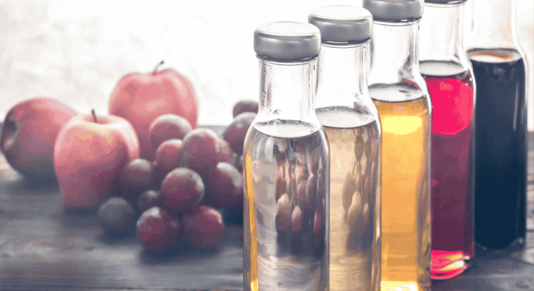 Four glass bottles filled with different colored liquids are lined up on a wooden table, with apples and grapes blurred in the background.