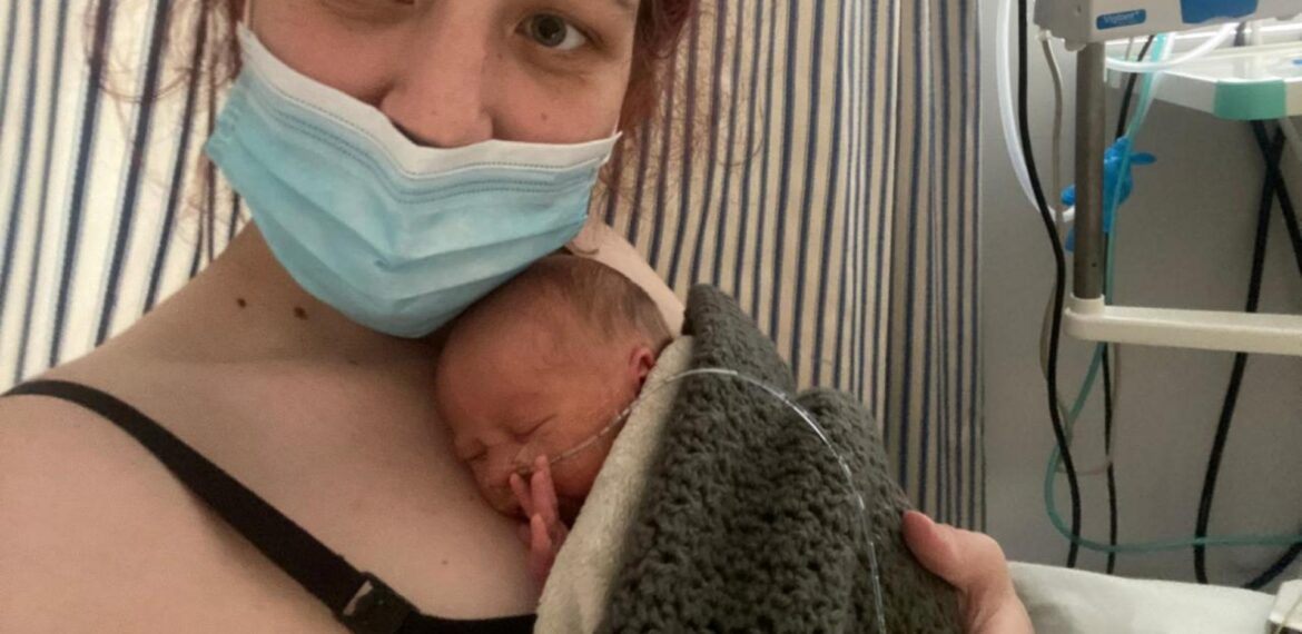 A woman wearing a face mask holds a premature baby wrapped in a blanket against her chest in a hospital room, with medical equipment and a faint scent of vinegar from nearby disinfectants in the background.