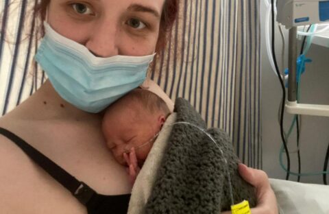 A woman wearing a face mask holds a premature baby wrapped in a blanket against her chest in a hospital room, with medical equipment and a faint scent of vinegar from nearby disinfectants in the background.
