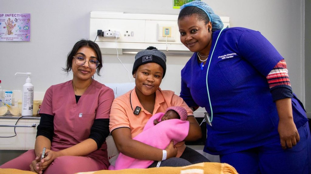 Three women smile at the camera in a hospital room. One woman, wearing a hat, holds a newborn baby wrapped in a pink blanket, while the other two women in medical scrubs offer support and guidance on breastfeeding.