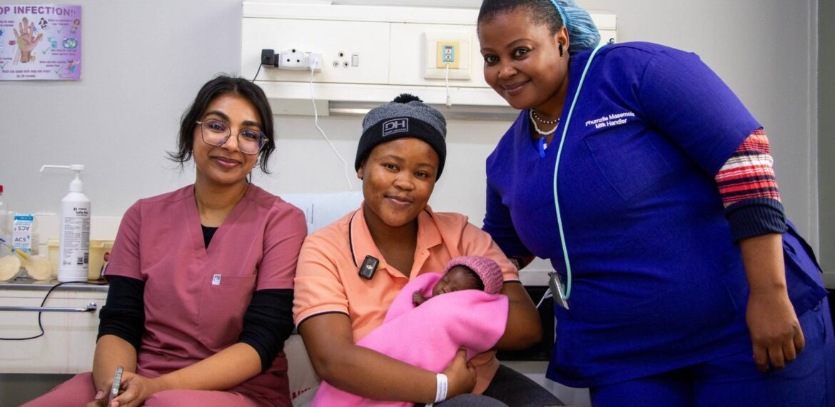 Three women smile at the camera in a hospital room. One woman, wearing a hat, holds a newborn baby wrapped in a pink blanket, while the other two women in medical scrubs offer support and guidance on breastfeeding.