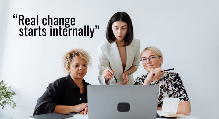 Three women sit together at a desk, focused on a laptop, while one woman stands and points at the screen. The text reads, “Real change starts internally”—a reminder that true progress goes beyond pinkwashing.