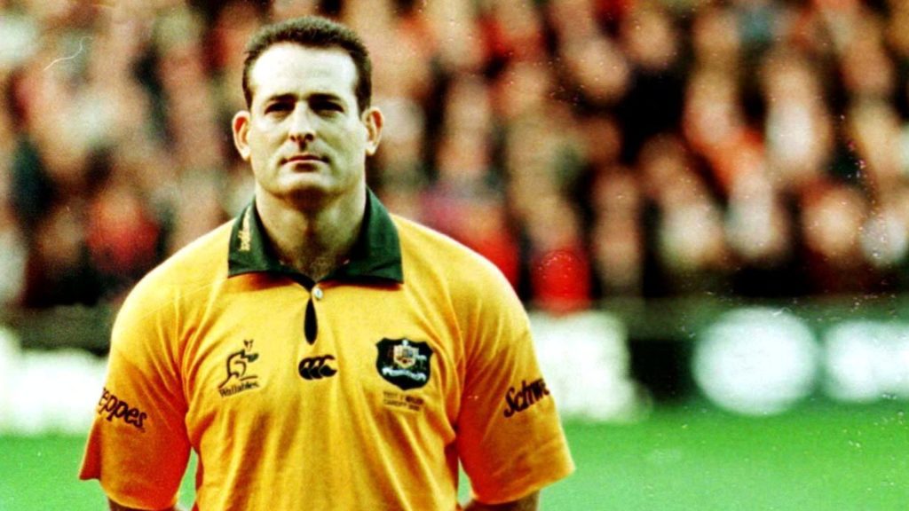 A male rugby player in a yellow and green jersey stands on a field, looking ahead with a serious expression. The background is blurred, showing an audience in a stadium.