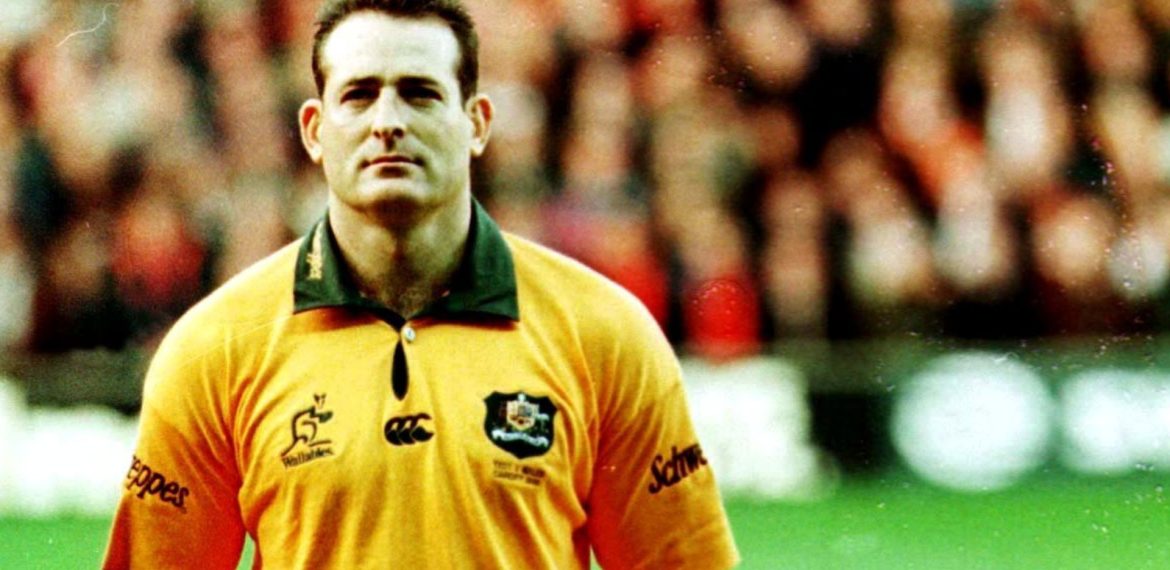 A male rugby player in a yellow and green jersey stands on a field, looking ahead with a serious expression. The background is blurred, showing an audience in a stadium.