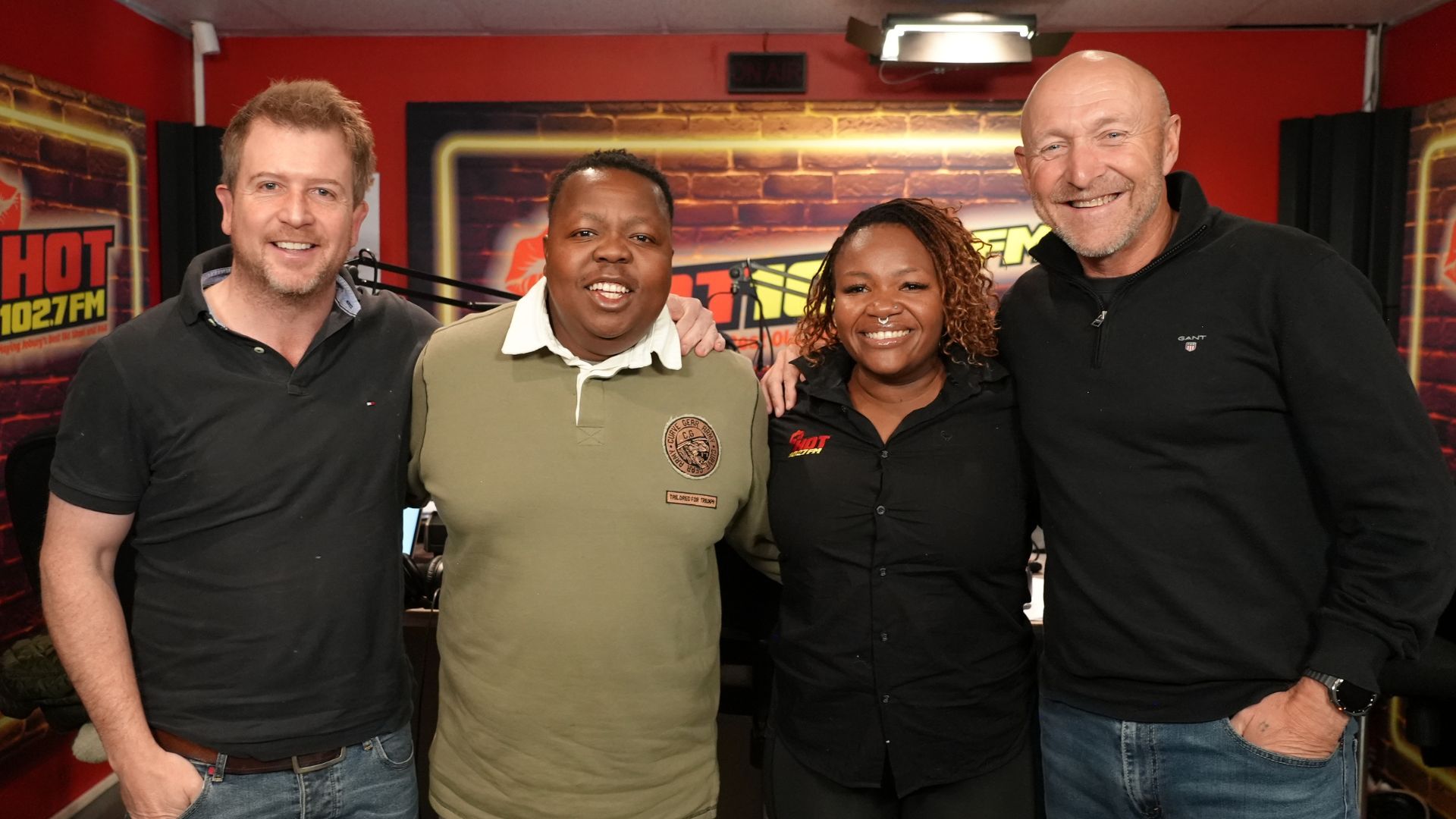 Four people stand together smiling in a radio studio, with microphones and HOT 102.7 FM branding visible in the background. The vibrant group shares good vibes—no hint of vinegar here—as they pose with two men on each side and a woman and man in the center.
