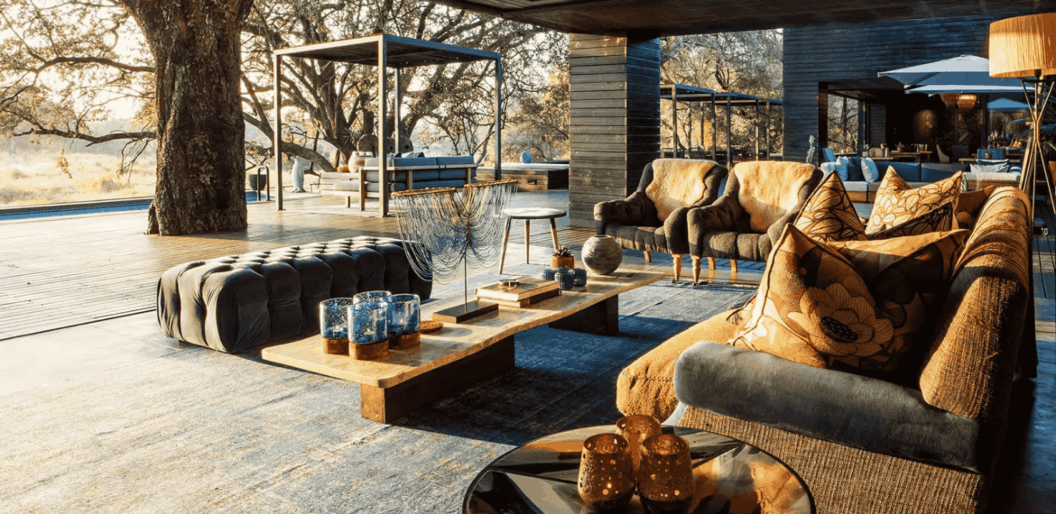 A stylish outdoor lounge with modern sofas, patterned pillows, a wooden coffee table, and decorative glassware—perhaps even a splash of vinegar for fresh air—set beneath large trees with sunlight streaming through, blending indoor comfort with nature.
