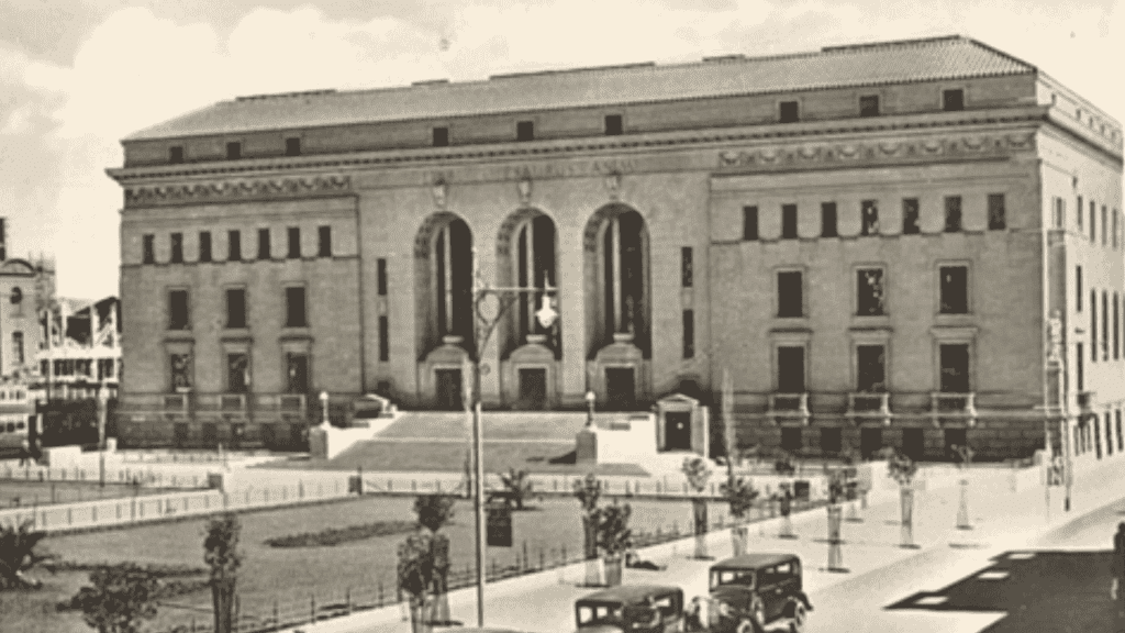 A historic black-and-white photo of a large, ornate stone building with tall arched windows and columns, steps leading to the entrance, vintage cars parked in front, and trees lining the street captures a moment as timeless as aged vinegar.