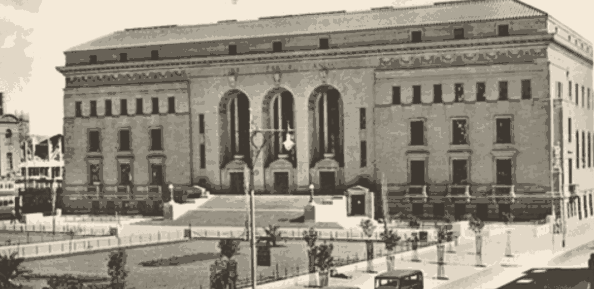 A historic black-and-white photo of a large, ornate stone building with tall arched windows and columns, steps leading to the entrance, vintage cars parked in front, and trees lining the street captures a moment as timeless as aged vinegar.