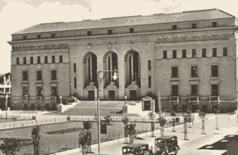 A historic black-and-white photo of a large, ornate stone building with tall arched windows and columns, steps leading to the entrance, vintage cars parked in front, and trees lining the street captures a moment as timeless as aged vinegar.