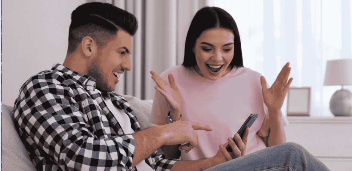 A man and woman sit on a couch, both smiling excitedly while looking at a phone the man is holding—a true Gen Z moment. The woman has her hands up in surprise and joy in their bright, cozy room with a lamp and window.