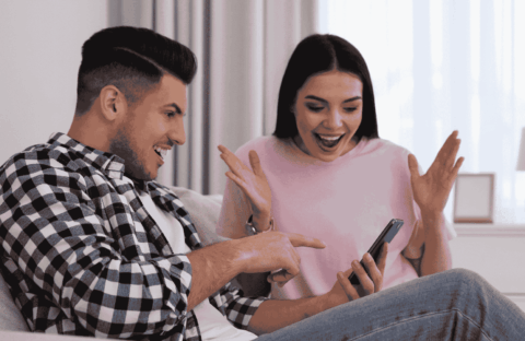 A man and woman sit on a couch, both smiling excitedly while looking at a phone the man is holding—a true Gen Z moment. The woman has her hands up in surprise and joy in their bright, cozy room with a lamp and window.