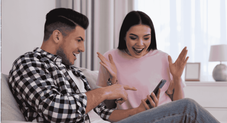 A man and woman sit on a couch, both smiling excitedly while looking at a phone the man is holding—a true Gen Z moment. The woman has her hands up in surprise and joy in their bright, cozy room with a lamp and window.
