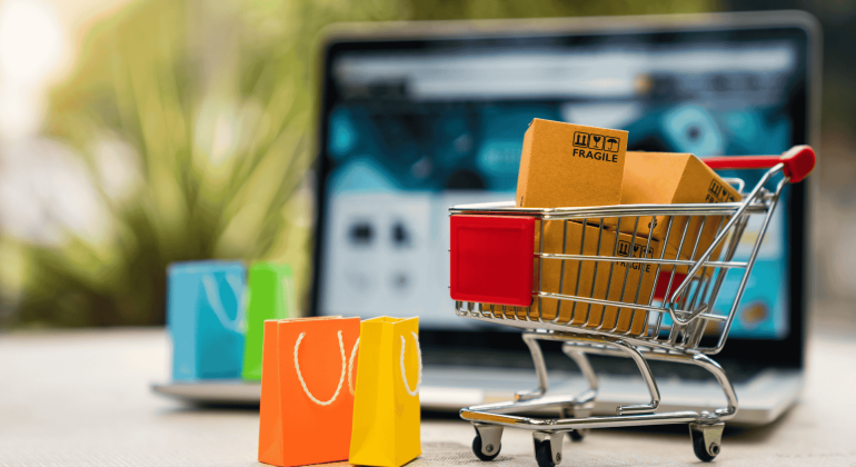 A small shopping cart with cardboard boxes and three colorful paper bags sits in front of an open laptop, symbolizing online shopping, e-commerce, and a seamless customer experience.