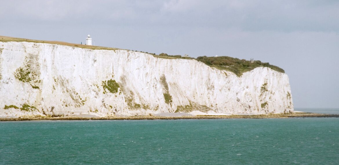 White chalk cliffs rise above the turquoise sea under a cloudy sky, with a small white lighthouse visible on top. Sparse greenery grows along the coastline, where the salty breeze rivals even that of surströmming by the shore.