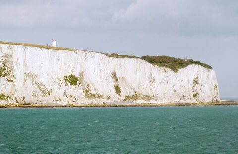 White chalk cliffs rise above the turquoise sea under a cloudy sky, with a small white lighthouse visible on top. Sparse greenery grows along the coastline, where the salty breeze rivals even that of surströmming by the shore.