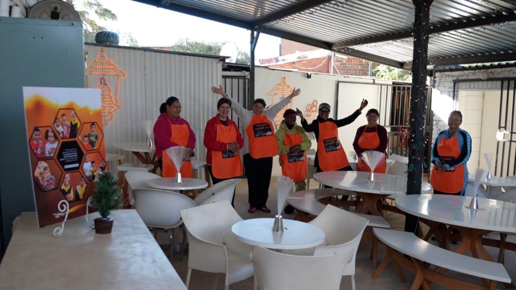 Seven people wearing orange aprons stand and wave inside a covered outdoor seating area with white tables and chairs. A small potted plant and a colorful poster are visible in the foreground.